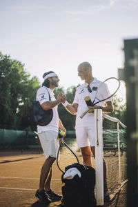 Men exchanging handshake holding tennis racket while standing at sports court