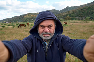 Happy, serious mature bearded man making a selfie on meadow with grazing horses. 