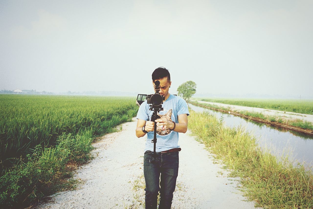 grass, lifestyles, field, casual clothing, rear view, tranquility, water, clear sky, tranquil scene, standing, leisure activity, full length, landscape, nature, copy space, rural scene, agriculture, sky