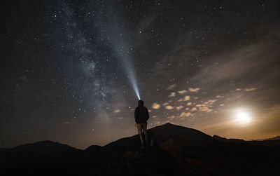 Rear view of man flashing light while standing on mountain against sky