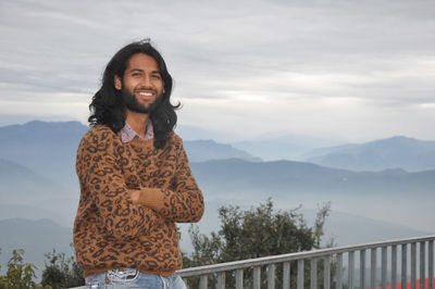 A smiling man with long hair looking at camera with crossed arms against the background of mountains