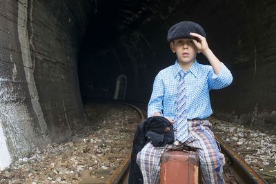 Portrait of boy sitting on suitcase at railroad track in tunnel 