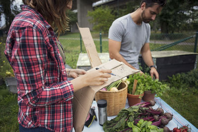 Midsection of woman writing on cardboard with man working in background at vegetable garden