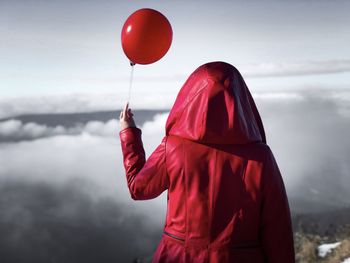 Rear view of woman wearing red hoodie holding a red balloon over the clouds