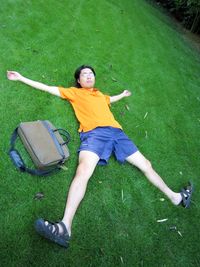 Young man with arms outstretched and legs apart lying by bag on grassy field