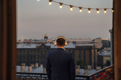 Male business person looking at buildings while listening music during sunset