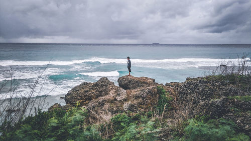 Man standing on rock by sea against cloudy sky