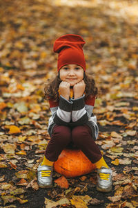 Happy cute little girl child in warm clothes sitting on a pumpkin in the autumn forest outdoors