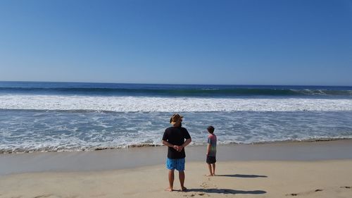 Father and son standing on sea shore at beach against clear blue sky