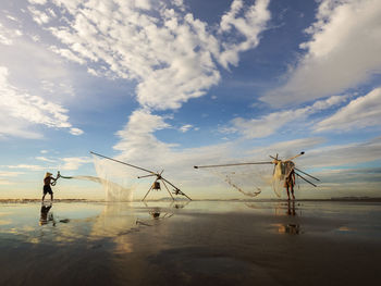 Fishermen with fishing nets standing at beach against sky during sunset