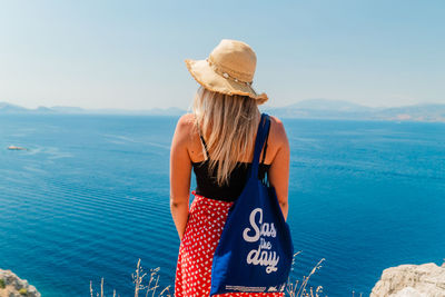 Rear view of a woman looking at blue the sea against sky with sun hat and tote bag.