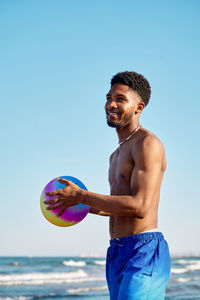 Positive african american male in shorts with colorful ball smiling and looking away while standing against waving sea and cloudless blue sky on sunny summer day on beach