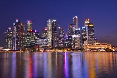 Illuminated modern buildings by river against sky at night