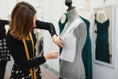 Anonymous woman with tape measure attaching paper cutout to mannequin with pin while making clothes in professional workshop