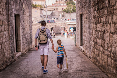 Rear view of father holding hands of son while walking in alley amidst buildings