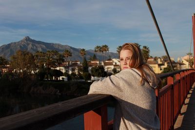 Thoughtful young woman leaning on bridge by mountain against sky