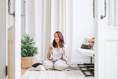 Young adult woman in cozy pajamas using mobile smartphone in room with christmas tree at home