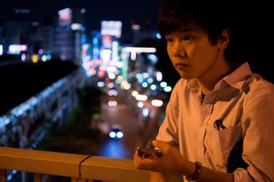 Thoughtful man using phone while standing at balcony in illuminated city during night