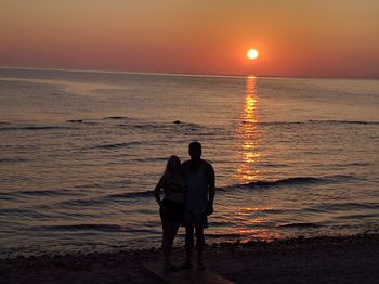 Full length of man and woman standing at beach against sky during sunset