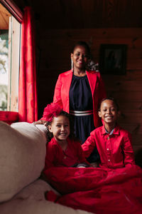 Cheerful young black female with cute laughing siblings in red clothes looking at camera while spending time together in cozy country house