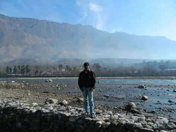 Portrait of man standing on stone wall at riverbank against mountains