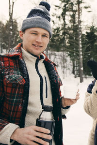 Portrait of smiling man having coffee while standing at park during winter