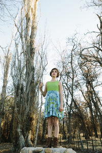 Low angle view of girl standing on rock amidst bare trees against sky in park