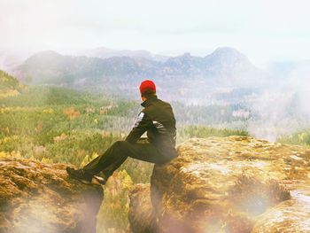 Man tourist sit on rocky cliff overlooking land below at misty border of horizon. abstract flare