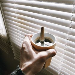 Cropped hand of man holding coffee against blinds