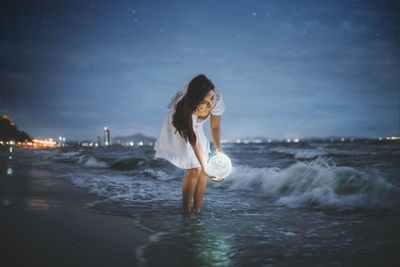 Side view of young woman standing in sea against sky at night