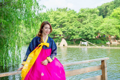 Portrait of smiling beautiful woman wearing hanbok while sitting on railing against lake