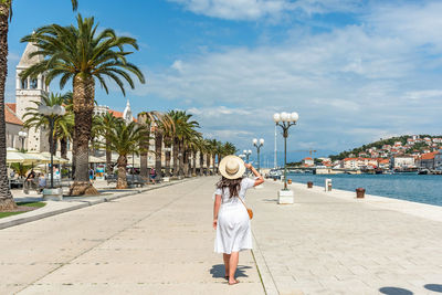 Rear view of woman wearing white sundress, walking on waterfront promenade in trogir, croatia