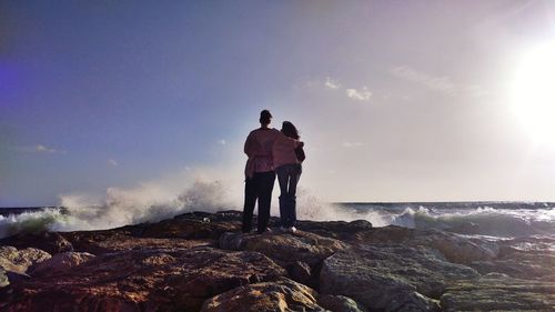 Rear view of friends standing at rocky shore against sky