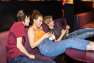 Happy multi-ethnic teenage friends relaxing on sofa while using smart phone at bowling alley