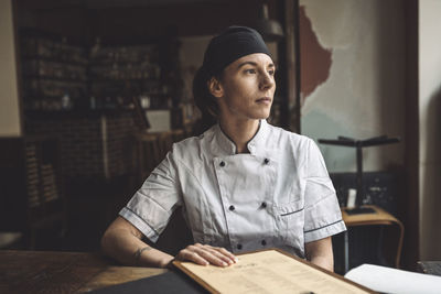 Female chef with menu looking away while sitting at restaurant