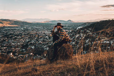 Male on rock posing in vintage clothes on golden hilltop overlooking a valley at sunset.
