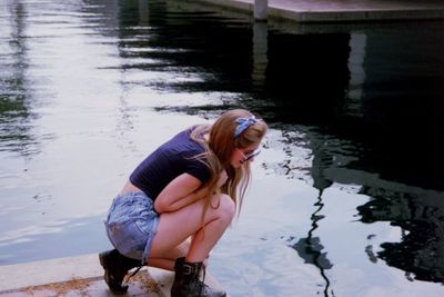 Side view of young woman looking at lake while crouching on pier