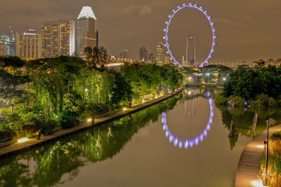 Illuminated ferris wheel by river against sky in city at night