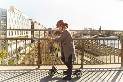 Full length side view of mature woman using smart phone while standing with electric push scooter on bridge in city