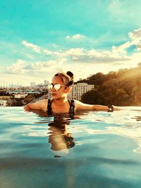 Young woman wearing sunglasses swimming in infinity pool against sky