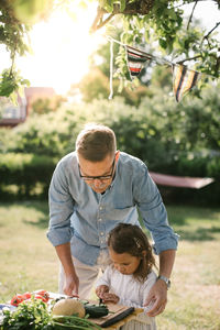 Grandfather assisting granddaughter in cutting vegetable at table in backyard