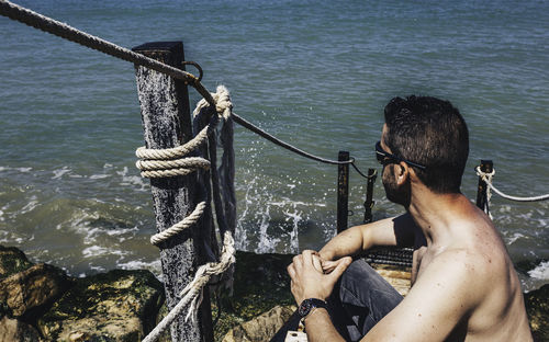 Shirtless man sitting by rope in sea