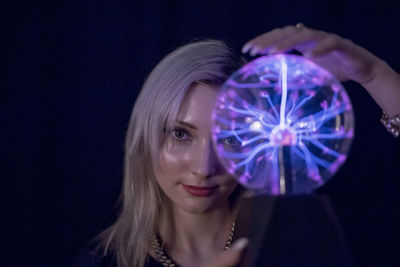 Close-up portrait of young woman holding plasma ball over black background