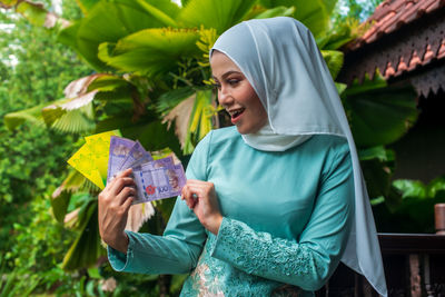 Smiling young muslim woman holding money and envelope while sitting outdoors during eid 