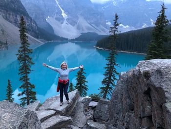 Portrait of happy woman with arms outstretched standing on cliff with lake and mountains in background