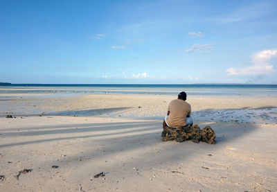 Rear view of man sitting on rock at beach against sky