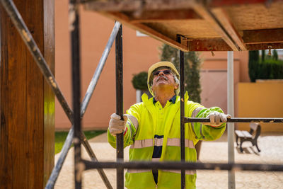 Positive skilled gray haired workman in uniform and protective helmet looking away while standing behind metal construction against building wall in sunny day