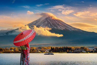 Woman wearing traditional clothing holding umbrella while standing by lake during sunset