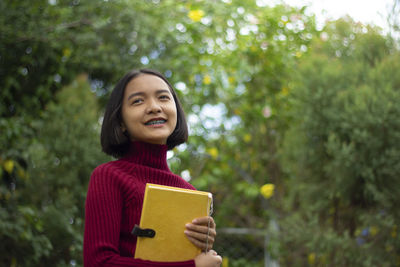 Girl holding book while standing against trees