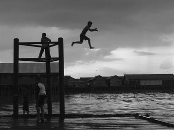 Silhouette person jumping in lake from diving platform against cloudy sky